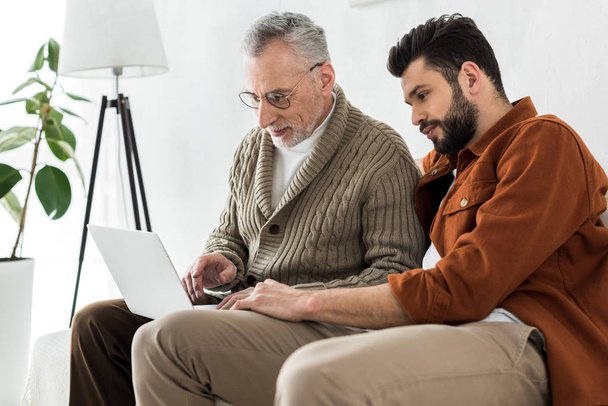 bearded man sitting with happy senior father and looking at laptop  - 写真・画像