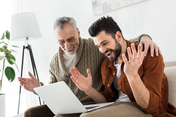 excited bearded man gesturing while sitting with senior father and looking at laptop  - Foto, Imagen