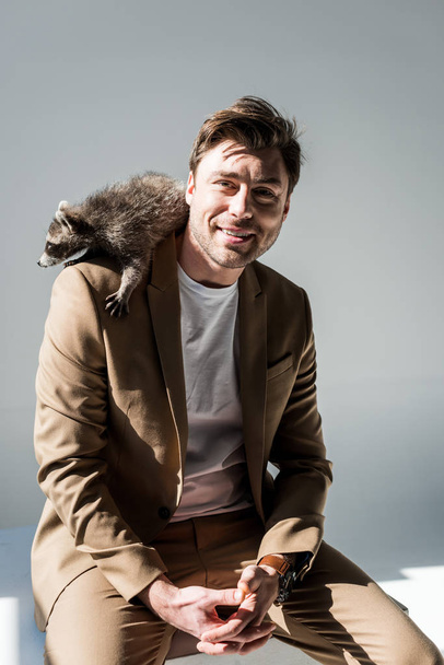 happy man in beige suite, with funny raccoon on shoulder, smiling and looking at camera on grey - Fotografie, Obrázek
