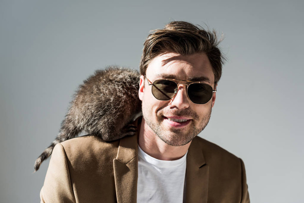smiling man in sunglasses with furry raccoon on shoulder on grey - Foto, Bild