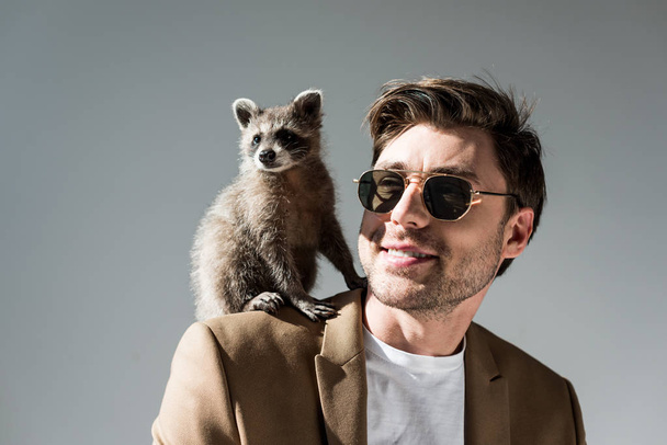 cheerful man in sunglasses with funny raccoon on shoulder on grey - Foto, Imagem