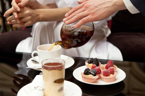 A man cares for a woman: pours her green tea. On the table are desserts: tiramisu and pastries with fresh berries. Without faces, in the frame of the hand. Closeup - Valokuva, kuva