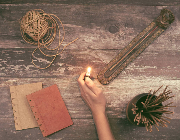 Woman hand lights a incense stick from a Buddha holder on a vintage natural wooden surface with books, hemp twine and many aromatic smelling incense sticks - Concept of spiritual ritual, creativity - Photo, Image