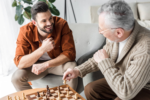 Pensionierter Vater mit Brille schaut glücklichen Sohn beim Schachspielen an  - Foto, Bild