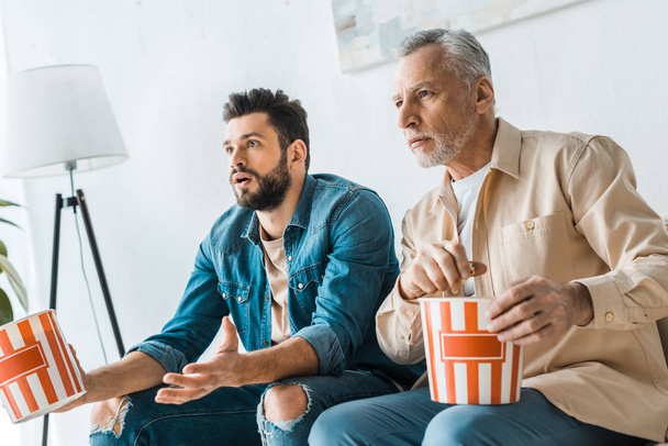 zaskoczony Starszy ojciec siedzi z synem przystojny i przytrzymując wiadro popcornu podczas oglądania telewizji - Zdjęcie, obraz