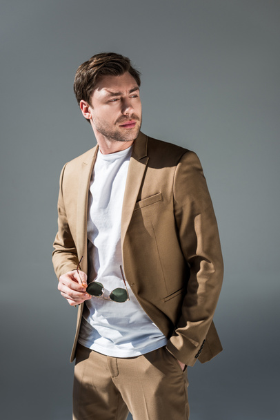 handsome man in beige suite holding sunglasses and looking away on grey - Photo, image