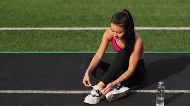 Urheilullinen aasialainen tyttö urheiluvaatteissa sitoo kengännauhoja lenkkarit istuu stadionilla ennen koulutusta
. - Materiaali, video