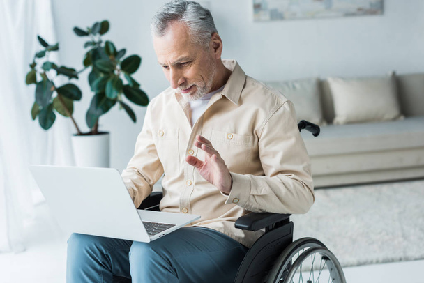 cheerful disabled retired man sitting in wheelchair and waving hand while having video call  - Fotoğraf, Görsel
