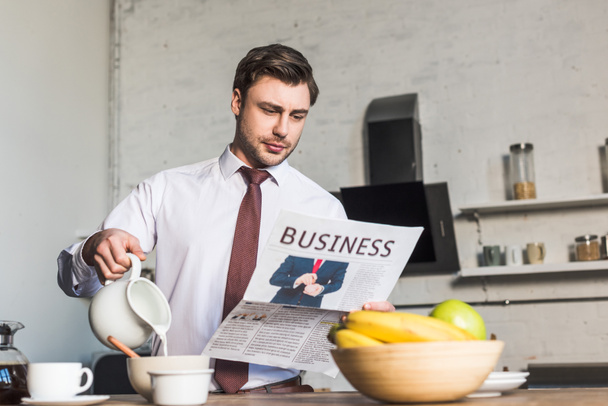 handsome man reading business newspaper while standing by kitchen table and pouring milk into bowl - Foto, afbeelding