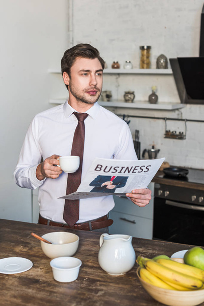 σίγουρος άνθρωπος κρατώντας κούπα καφέ και επιχειρηματική εφημερίδα, ενώ στέκεται στην κουζίνα στο σπίτι - Φωτογραφία, εικόνα