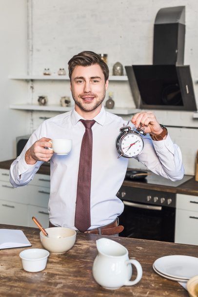 cheerful man holding coffee cup and alarm clock while standing in kitchen at home - Foto, Imagem