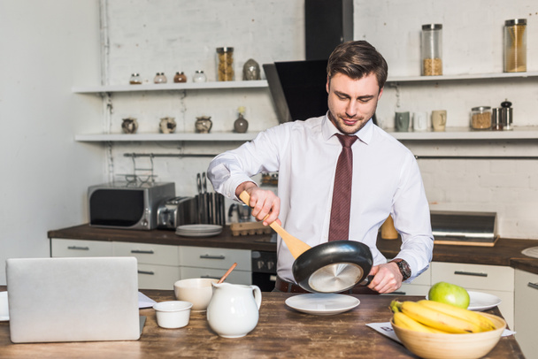 hombre guapo con sartén preparando el desayuno mientras está de pie junto a la mesa de la cocina
 - Foto, Imagen