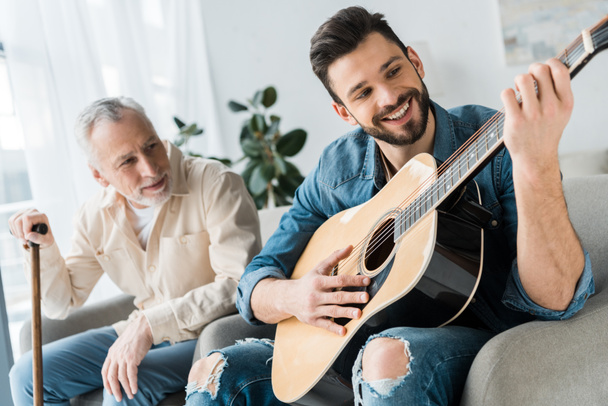 Fröhlicher bärtiger Mann spielt Akustikgitarre bei älterem Vater zu Hause  - Foto, Bild