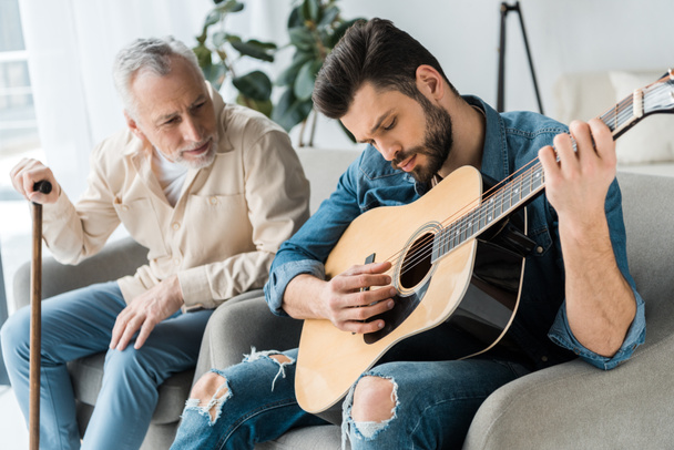 guapo barbudo hombre tocando la guitarra acústica cerca feliz padre mayor en casa
  - Foto, Imagen