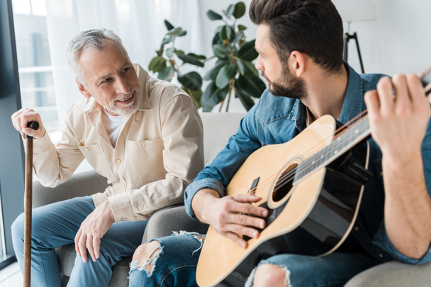 Bärtiger Sohn spielt Akustikgitarre bei glücklichem Senior-Vater zu Hause  - Foto, Bild
