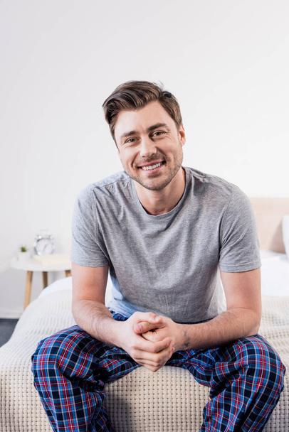 handsome cheerful man sitting on bedding at home and looking at camera - Фото, изображение