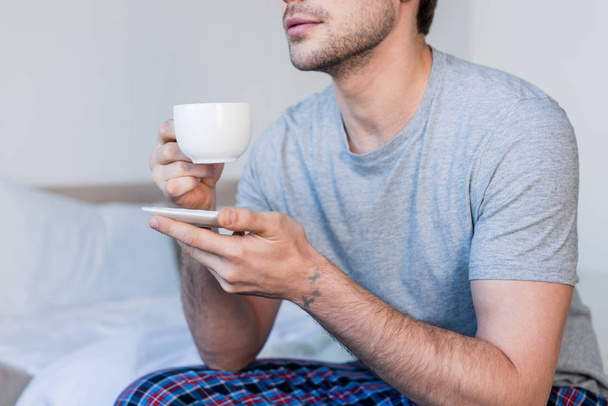 cropped view of man in grey t-shirt sitting on bed and holding coffee cup - Photo, Image