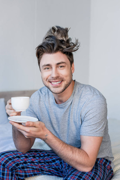 весёлый человек со смешным енотом на голове держит чашку кофе и смотрит в камеру
 - Фото, изображение