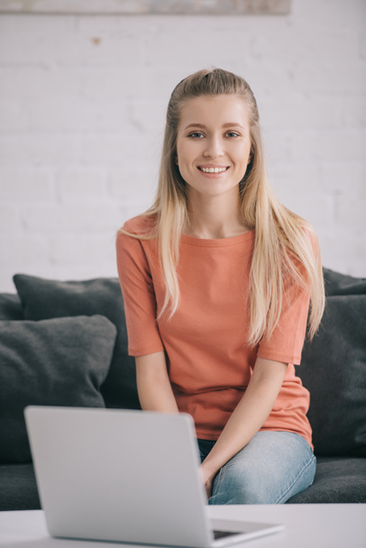 happy blonde freelancer sitting on sofa near laptop at home  - Φωτογραφία, εικόνα