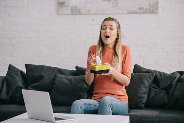 attractive woman sneezing while holding tissue box near laptop - Photo, Image