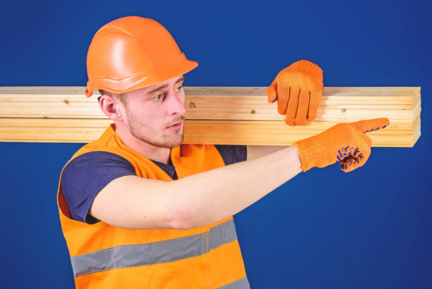 Carpenter, woodworker, strong builder on busy face carries wooden beam on shoulder. Man in helmet, hard hat and protective gloves pointing direction, blue background. Wooden materials concept - Foto, imagen