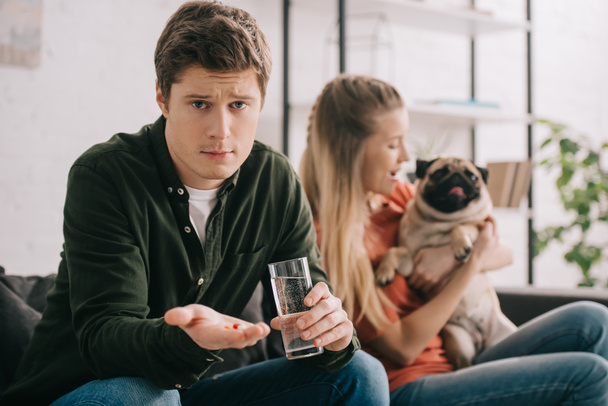selective focus of handsome man allergic to dog holding glass of water and pills near blonde woman with pug  - Fotografie, Obrázek