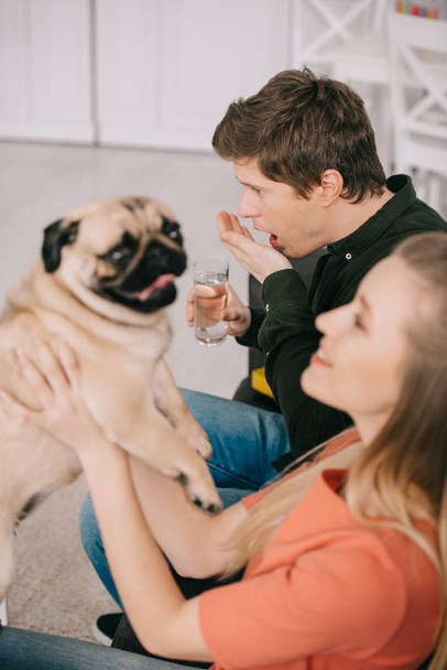 valikoiva painopiste komea mies allerginen koira tilalla lasi vettä ja ottaen pillereitä lähellä blondi nainen mopsi
  - Valokuva, kuva
