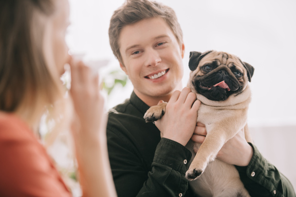 selective focus of cheerful handsome man holding pug and looking at woman allergic to dog with tissue  - Valokuva, kuva