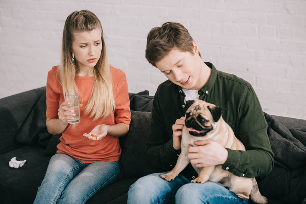 привлекательная блондинка с аллергией на собаку, держащую стакан воды и таблетки рядом с мужчиной с мопсом
  - Фото, изображение