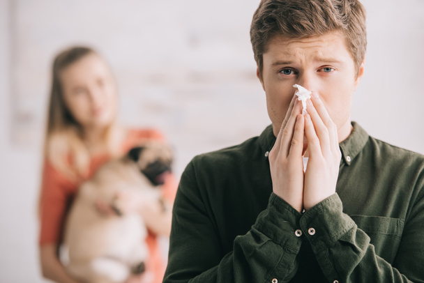 Selektywny fokus człowieka alergiczne psu kichanie w tkanki i patrząc na kamery w pobliżu kobieta z Mops  - Zdjęcie, obraz
