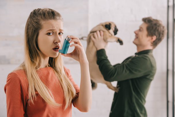 Selektivní fokus rozrušená žena alergický na psa Držte inhalátor a při pohledu na fotoaparát blízko člověka s Mops  - Fotografie, Obrázek