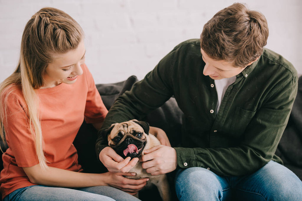 happy blonde woman and cheerful man touching cute pug dog on sofa  - Zdjęcie, obraz