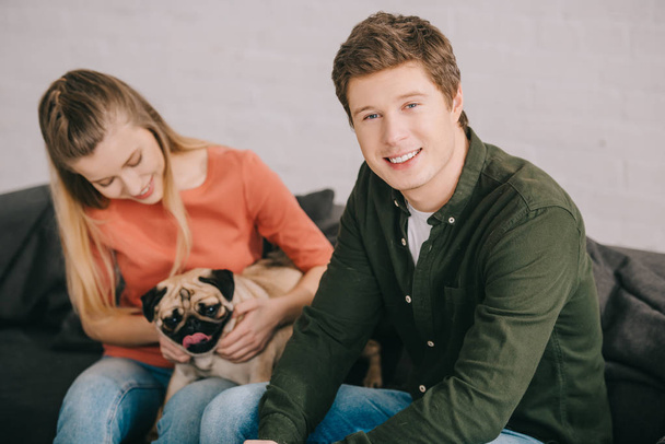 valikoiva painopiste komea hymyilevä mies istuu lähellä naista ihana mopsi koira
  - Valokuva, kuva