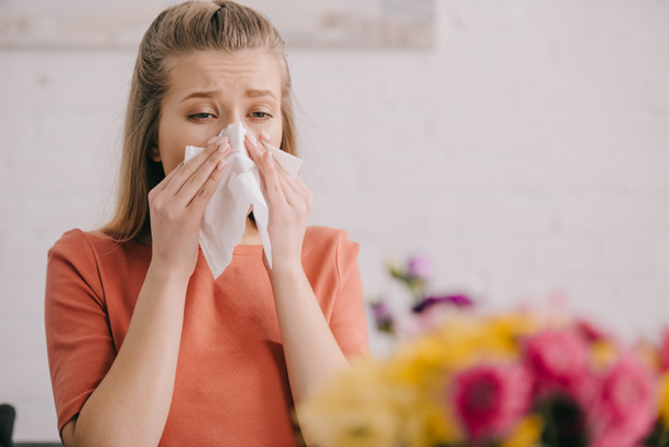 selective focus of blonde woman looking at flowers and sneezing in tissue while having pollen allergy - Fotó, kép