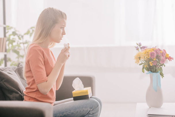 side view of sad blonde woman with pollen allergy holding tissue and looking at flowers in vase  - Photo, Image