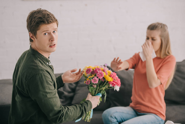 upset man holding flowers and looking at camera near blonde woman with pollen allergy sneezing in tissue  - Photo, Image