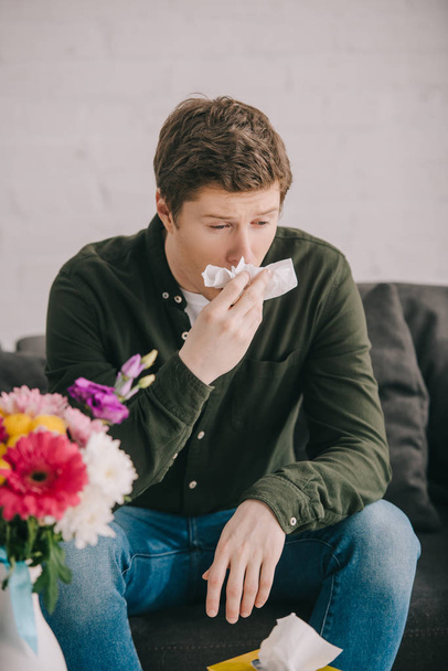красивый мужчина с аллергией на пыльцу чихает рядом с цветами, сидя на диване
  - Фото, изображение