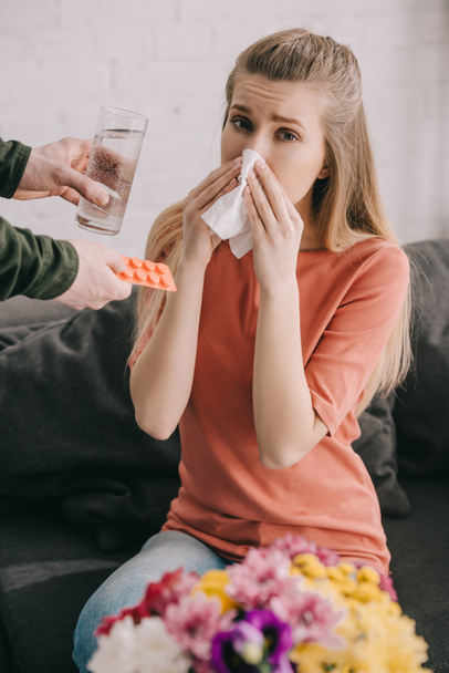 cropped view of man holding glass of water and pills near woman with pollen allergy sneezing in tissue near flowers  - Foto, afbeelding