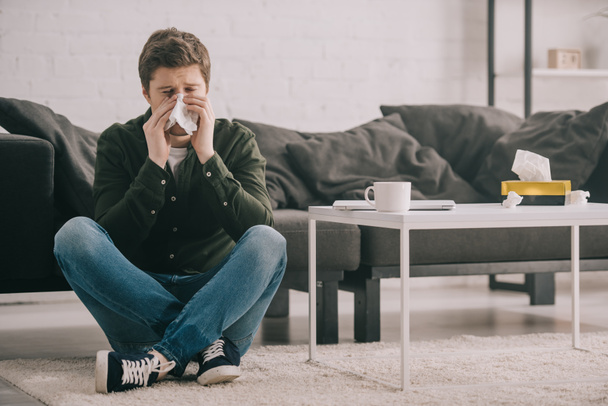 man sitting on carpet with crossed legs and sneezing in tissue near coffee table with cup  - Foto, Imagem