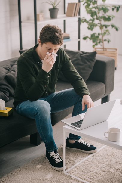 hombre enfermo estornudando en el tejido mientras está sentado en el sofá y el uso de la computadora portátil en casa
  - Foto, Imagen