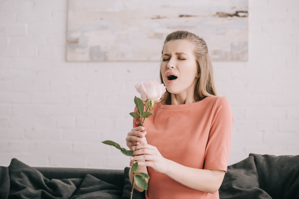 atractiva chica rubia con alergia al polen estornudos mientras sostiene rosa en casa
  - Foto, imagen
