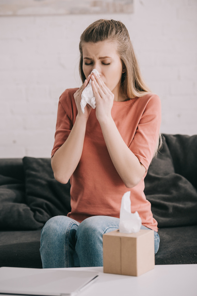 attractive blonde girl sneezing while holding tissue near tissue box at home  - Foto, imagen