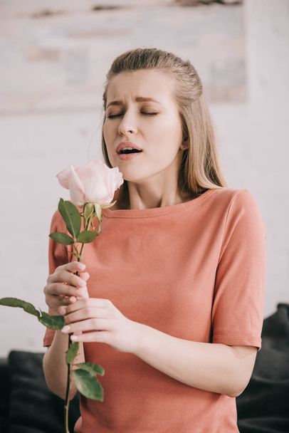 beautiful blonde girl with pollen allergy sneezing with closed eyes while holding rose at home  - Photo, Image
