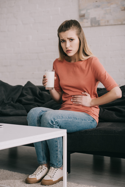 upset blonde woman with lactose intolerance holding glass of milk while sitting on sofa  - Photo, Image