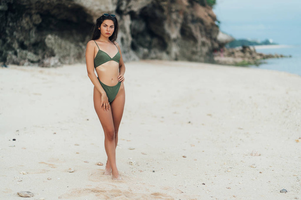 Beautiful tanned girl in green swimwear and sunglasses posing on beach background of rock - 写真・画像