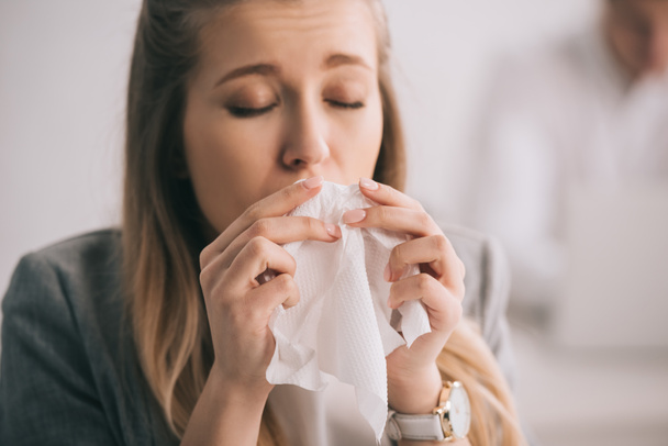 blonde businesswoman sneezing in tissue with closed eyes in office  - Foto, Imagen