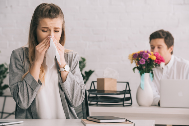 blonde businesswoman with pollen allergy sneezing in tissue near coworker smelling flowers in office  - 写真・画像