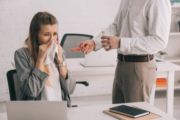 cropped view of businessman holding pills and glass of water near blonde colleague sneezing in tissue  - Photo, Image