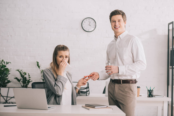 happy businessman holding pills and glass of water near blonde colleague in office  - Foto, Bild