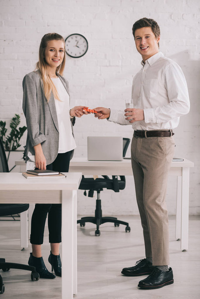 cheerful businessman holding pills and glass of water near happy blonde colleague in office  - Foto, afbeelding